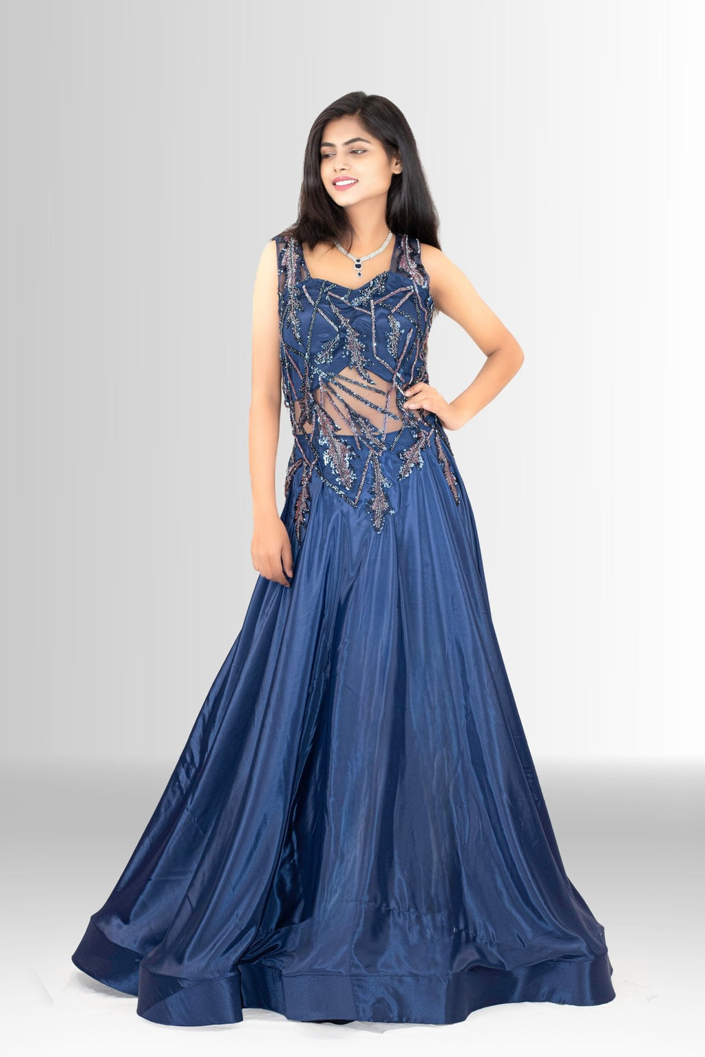 ull shot of a woman smiling, wearing a navy blue A-line gown with an intricate sequin and embroidered sheer illusion bodice, standing on a white background.