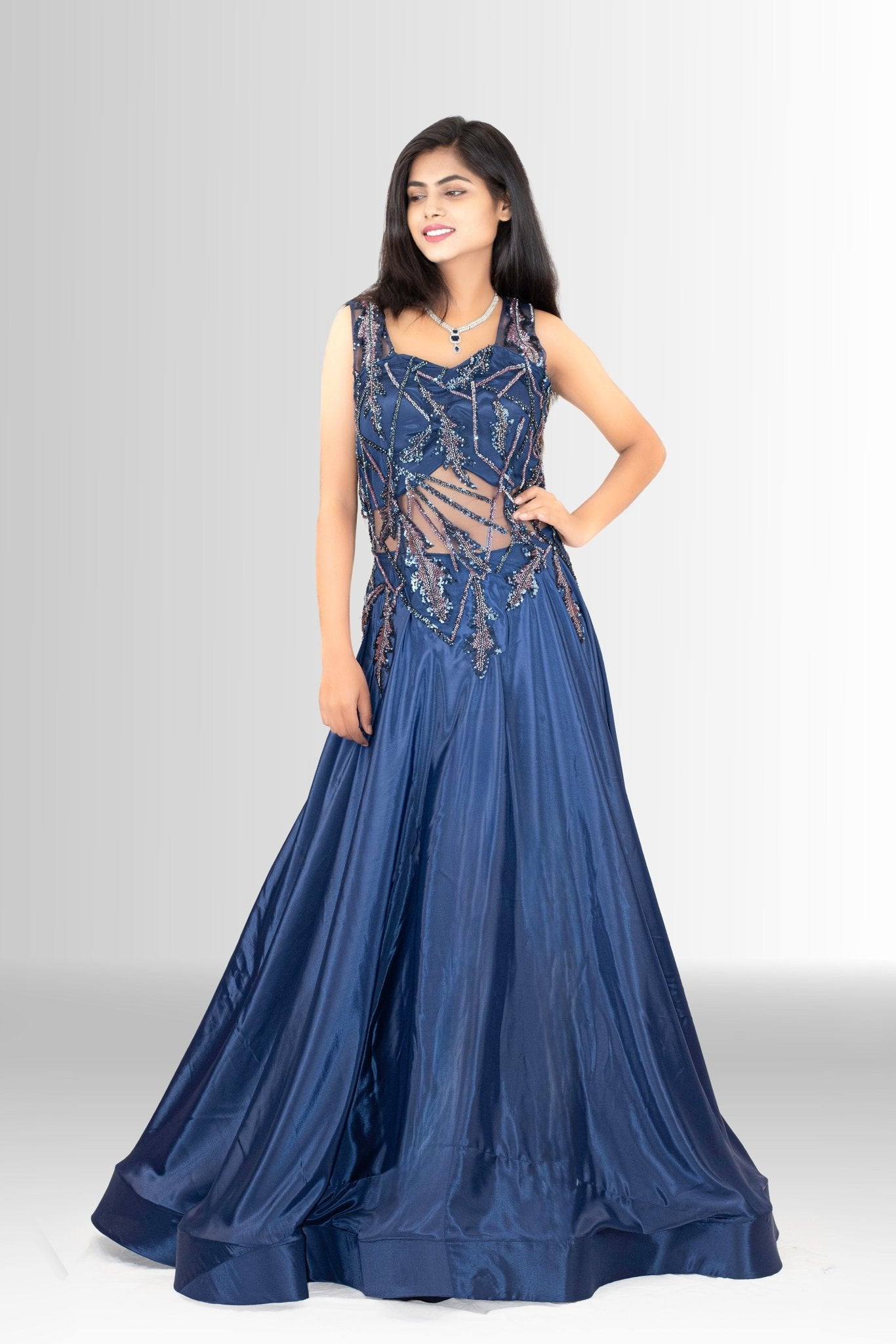 ull shot of a woman smiling, wearing a navy blue A-line gown with an intricate sequin and embroidered sheer illusion bodice, standing on a white background.